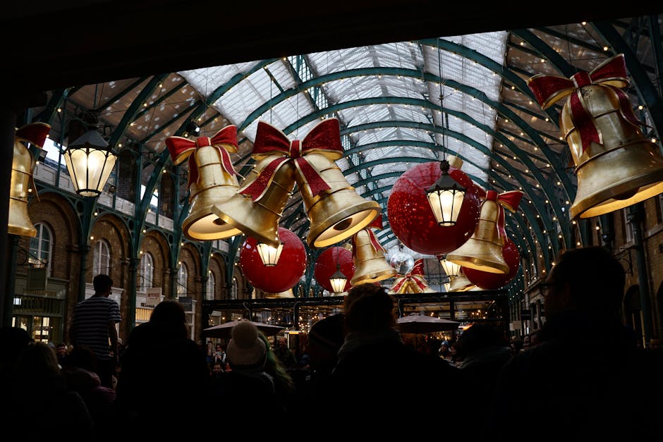 Interior view of a shopping arcade or indoor market with a high, curved glass ceiling decorated for the holiday season. Large gold and red Christmas ornaments styled as bells with red ribbons, along w