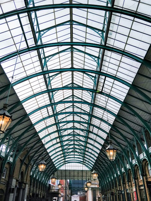 Interior view of a shopping arcade or indoor market with a high, curved glass ceiling decorated for the holiday season. Large gold and red Christmas ornaments styled as bells with red ribbons, along w