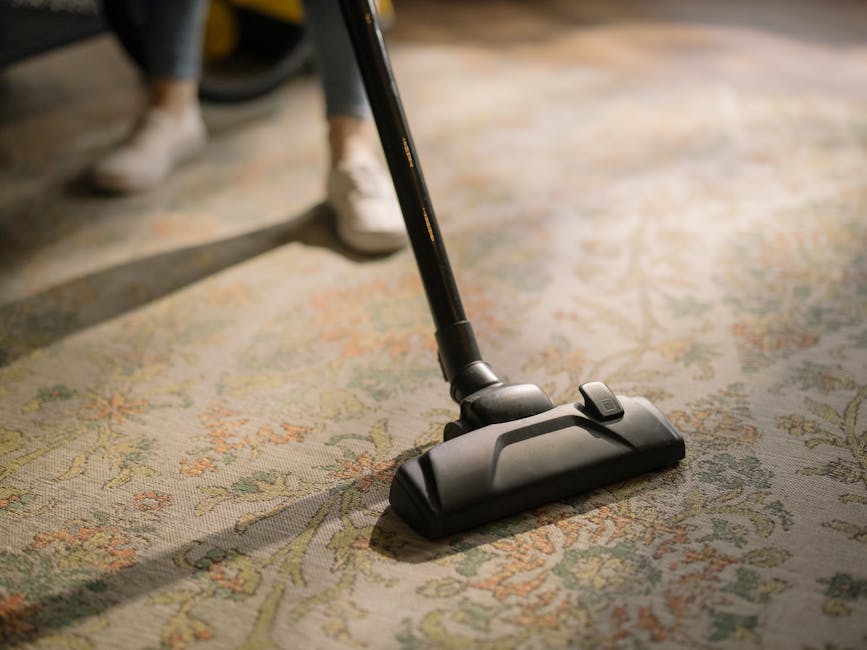 A person kneeling on a patterned area rug, using a yellow portable vacuum cleaner for surface cleaning. The individual has light brown, curly hair and is wearing a beige long-sleeve shirt and blue jea